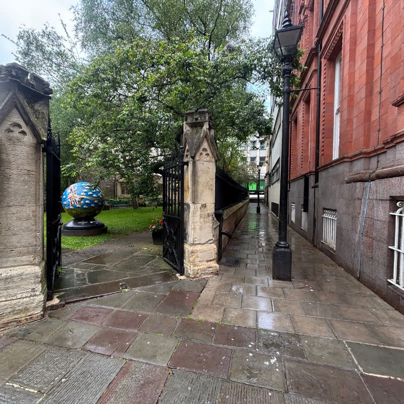 St Stephens Church Gardens Social Story Picture 2 - A photo of a wide gate with stone pillars on either side. The path is paved leading into the gate and the gardens beyond. There is a narrow paved path to the right of the gate which runs alongside a red brick building. It has been raining and so the ground is shiny and wet.