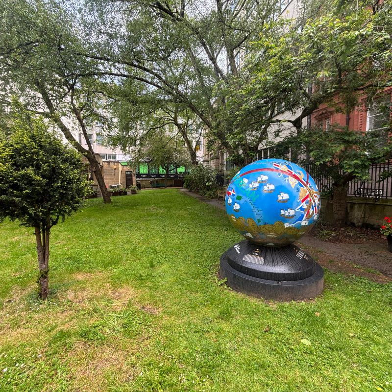 St Stephens Church Gardens Social Story Picture 3 - A photo a grassy area with a large blue ball scultpture. The sculpture is on a black plinth and has colourful drawings on. There are tall green leafy trees behind it.