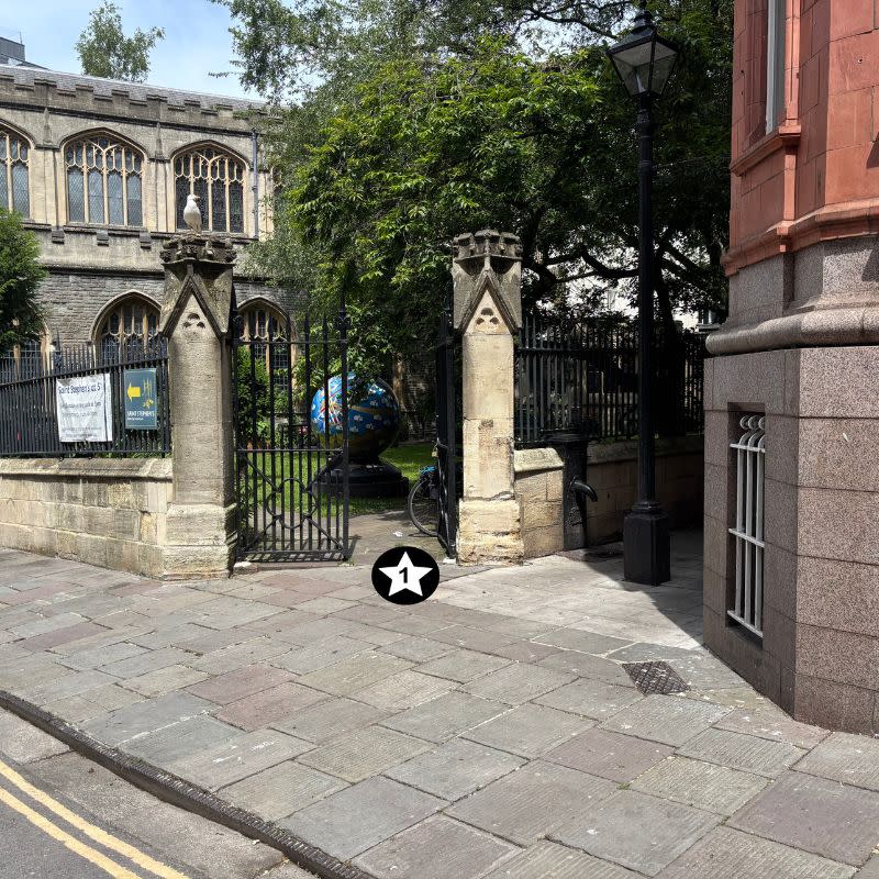 St Stephens Church Gardens Social Story Picture 3 - A photo of a wide gate with stone pillars on either side. The path is paved leading into the gate and the gardens beyond. There is a narrow paved path to the right of the gate which runs alongside a red brick building. It has been raining and so the ground is shiny and wet.