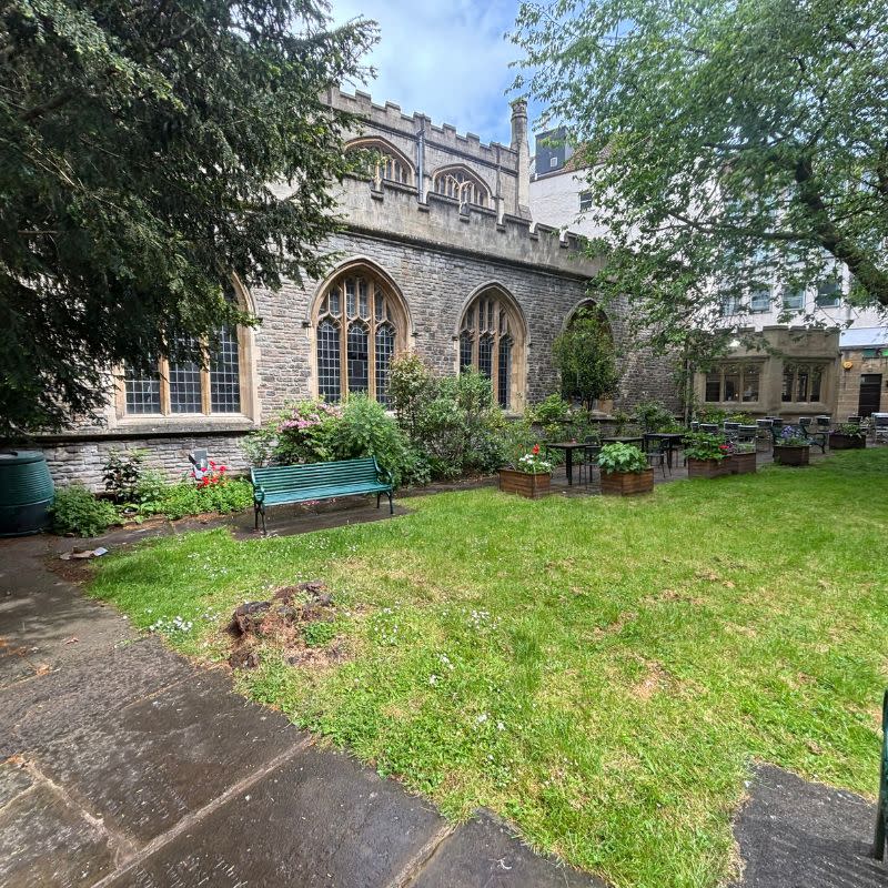 St Stephens Church Gardens Social Story Picture 4 - A photo a grassy lawn area surrounded by narrow paved paths. There is a church behind the path and benches and flowers pots on the path running alongside the church.