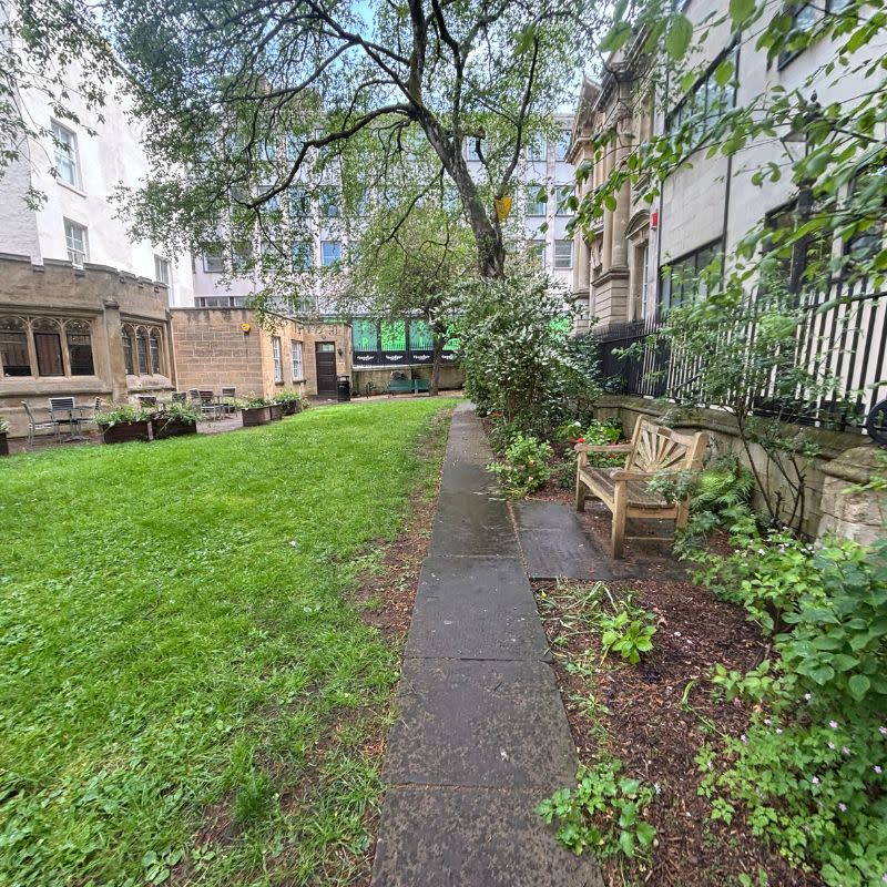 St Stephens Church Gardens Social Story Picture 5 - A photo of a narrow paved path running alongside a grassy lawn area to the left of the path. On the right is a flower bed, a wooden bench and a black metal fence on top of a stone wall.