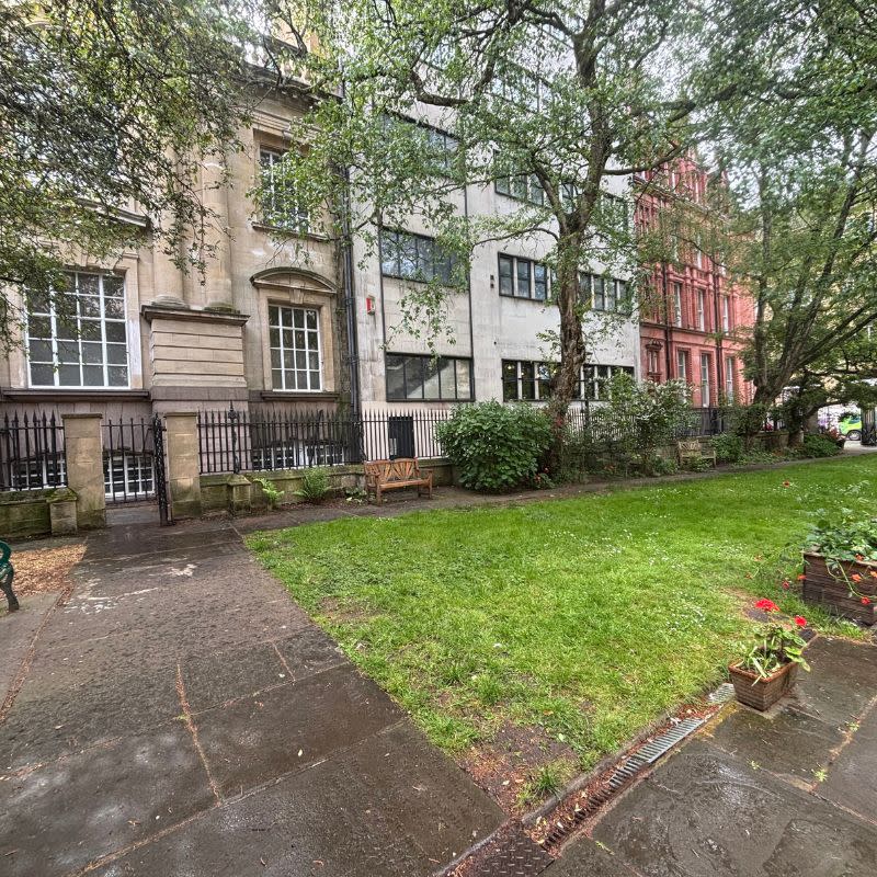 St Stephens Church Gardens Social Story Picture 6 - A photo of a church garden with a lawn area in the middle, a paved path to the left and a stone wall with a black metal fence running along the top. There is a gap where a metal gate is located with stone pillars on either side.