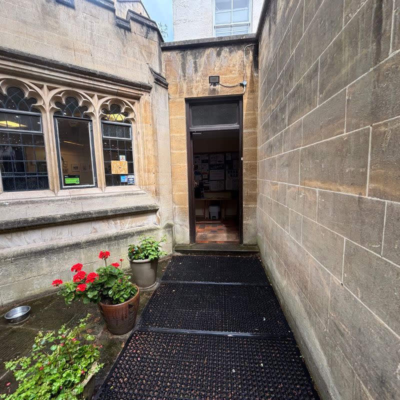 St Stephens Church Gardens Social Story Picture 7 - A photo of an entrance and doorway. The building is made of stone and there are flowerpots with red flowers running alongside the entrance path on the left side. The door is black.