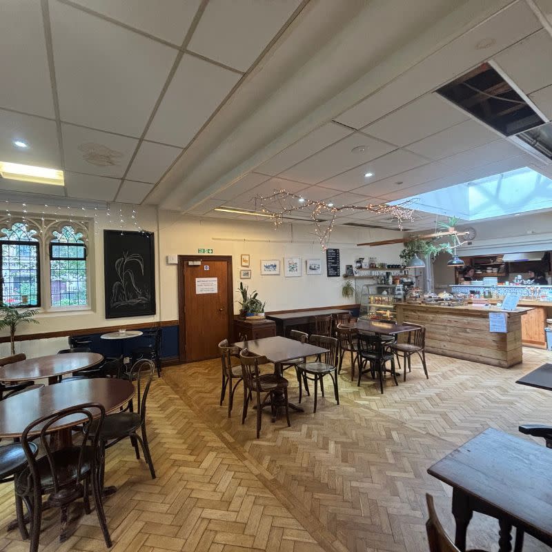 St Stephens Church Gardens Social Story Picture 8 - A photo of an empty cafe. There are 5 wooden tables with chairs, a long wooden counter at the back with food on display. There is a white ceiling.
