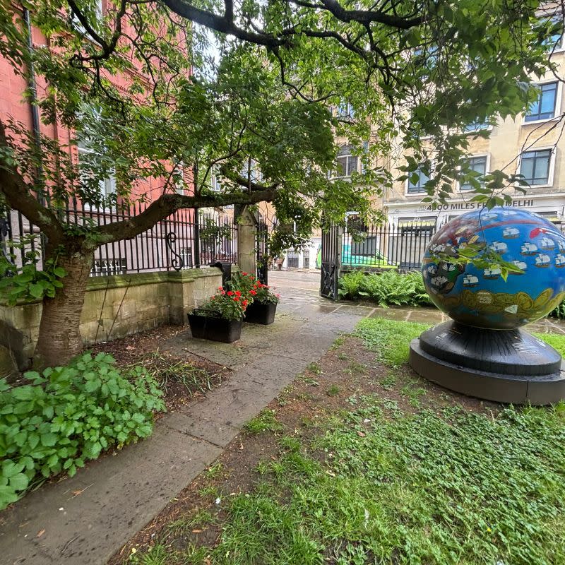 St Stephens Church Gardens Social Story Picture 9 - A photo of a narrow path with a tree and a flower bed on the left side. On the right of the path is a lawn area with a big blue ball sculpture on. There is a gate at the end of the path leading out of the garden.