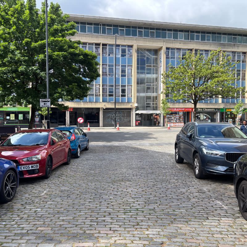 Temple Church Gardens Social Story Picture 12 - A photo of a cobbled street with parked cars lining each side. The cobbled street leads out to a wider busier road. There is a tall, wide building on the other side of the road ahead.