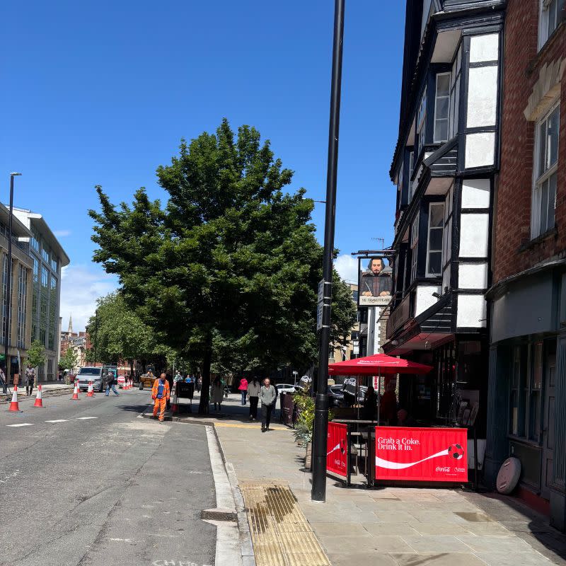Temple Church Gardens Social Story Picture 1 - A photo of a pavement on the right with a paved road on the left. There is a pub ahead with a low red barrier around some outside seating. There is a large tree up ahead on the pavement past the pub.