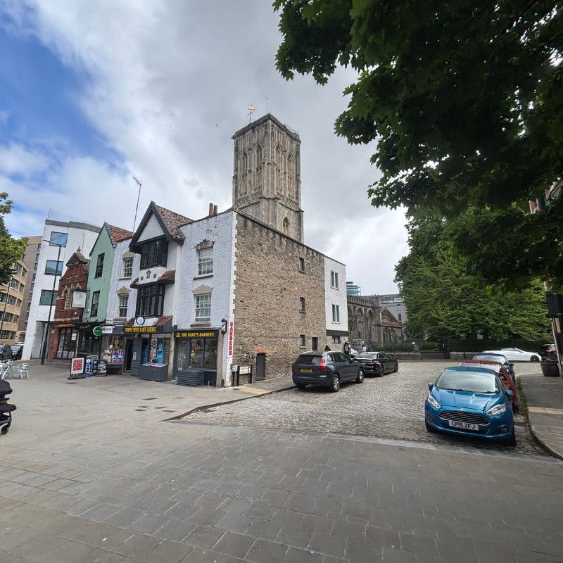 Temple Church Gardens Social Story Picture 2 - A photo of a wide path leading up to a quiet road with parked cards. There are some three storey building along the left side of the road and a wonky church tower begind them.