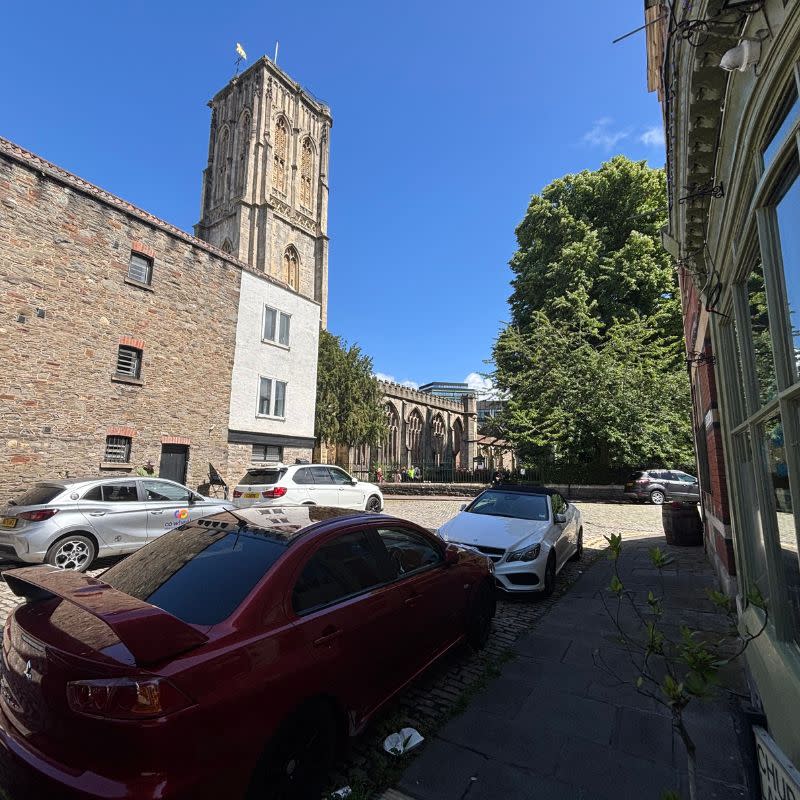 Temple Church Gardens Social Story Picture 2 - A photo of a wide path leading up to a quiet road with parked cards. There are some three storey building along the left side of the road and a wonky church tower begind them.