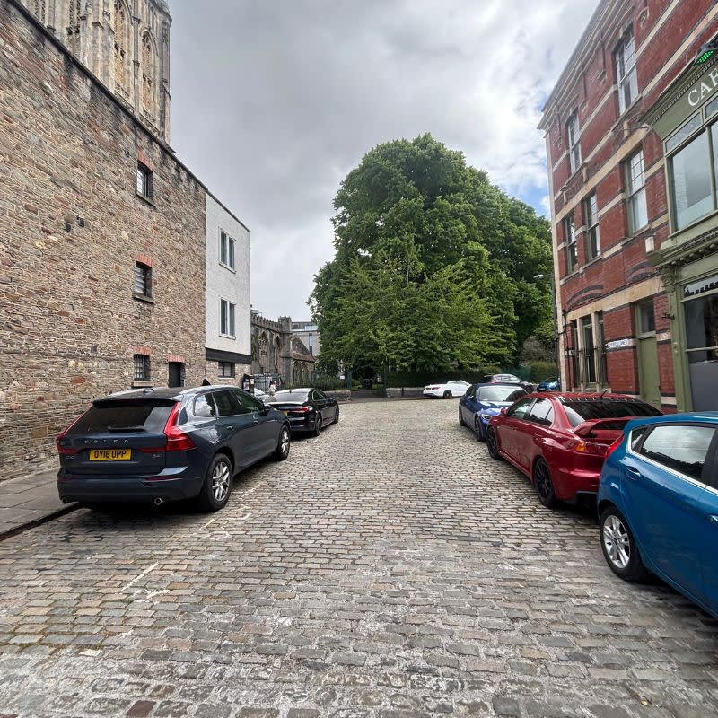 Temple Church Gardens Social Story Picture 3 - A photo of a cobbled street with building and cars parked on either side. Up ahead there is a large mature tree.