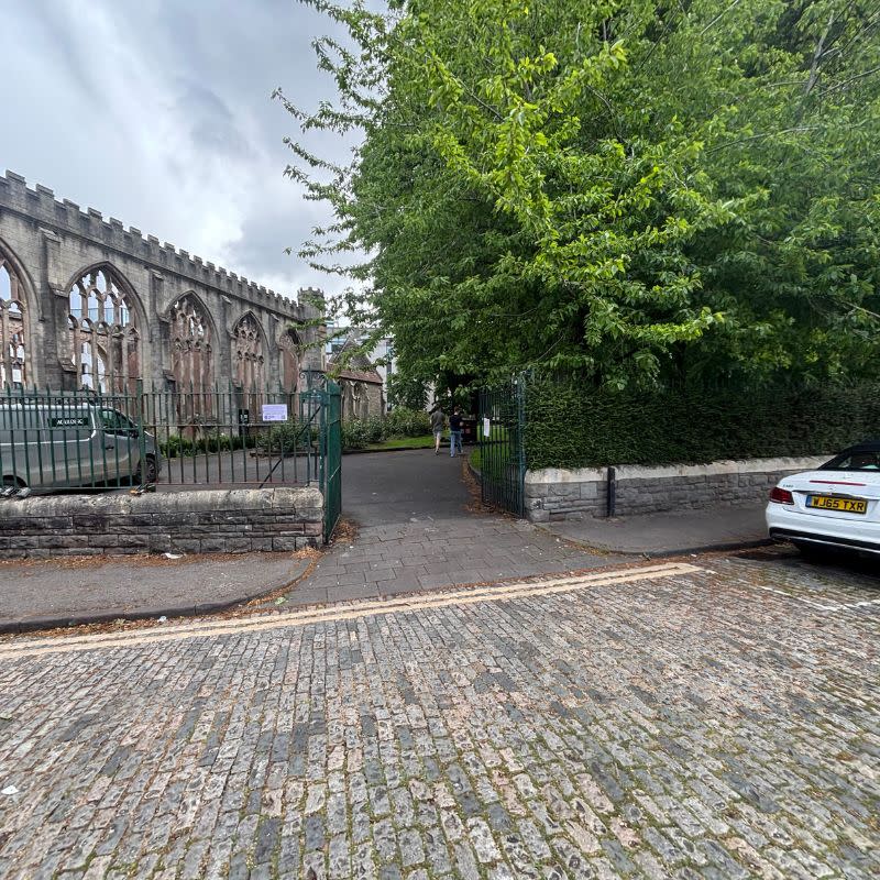 Temple Church Gardens Social Story Picture 4 - A photo of a cobbled street in the foreground and a dark green gate ahead, which leads into a garden with a church ruin on the left and a large mature green tree to the right.