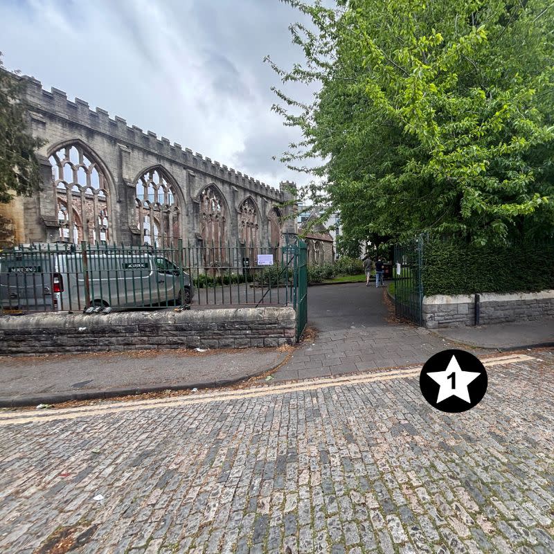 Temple Church Gardens Social Story Picture 4 - A photo of a cobbled street in the foreground and a dark green gate ahead, which leads into a garden with a church ruin on the left and a large mature green tree to the right.