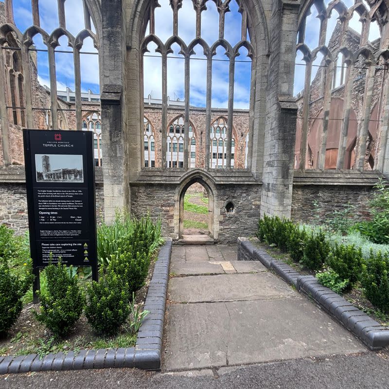 Temple Church Gardens Social Story Picture 7 - A photo of a entrance to a church ruin, there is a black information sign on the left side and a smaller arched doorway in a stone wall leading into the church. The windows of the church are just the stone structure without any glass and the church has no roof so is open to the sky.