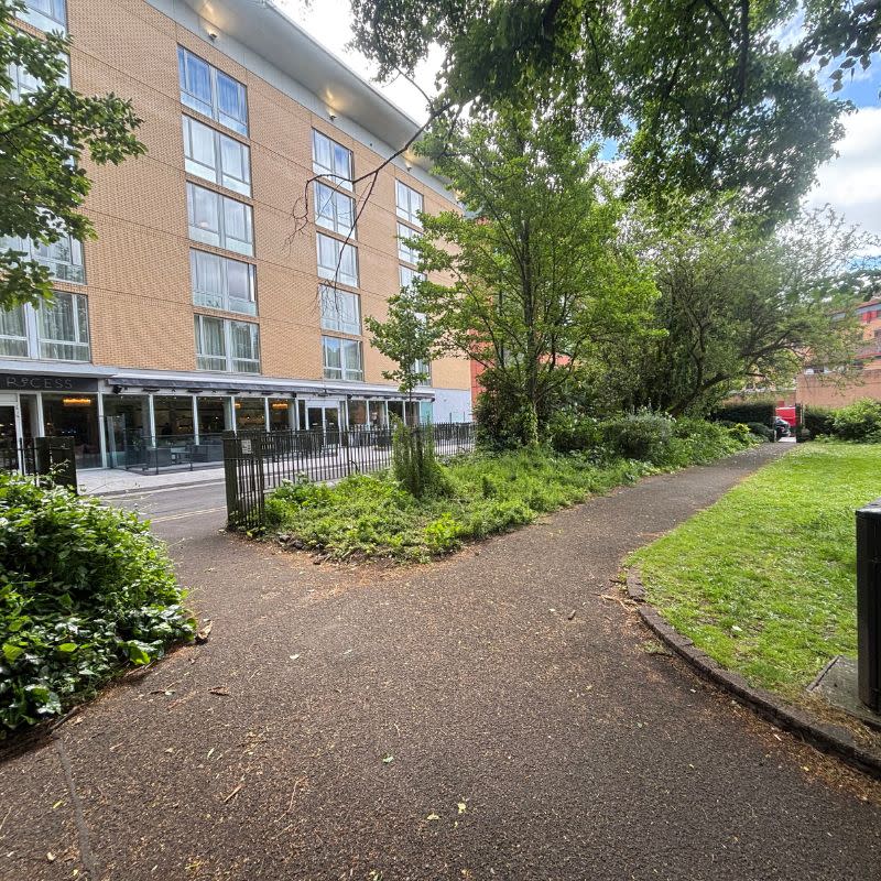 Temple Church Gardens Social Story Picture 9 - A photo of a path in a garden leading out to a wide, open gate through a green metal fence. There is a tall building, a hotel, on the other side of the gate. There are some bushes and small trees lining the fence alongside the gate.
