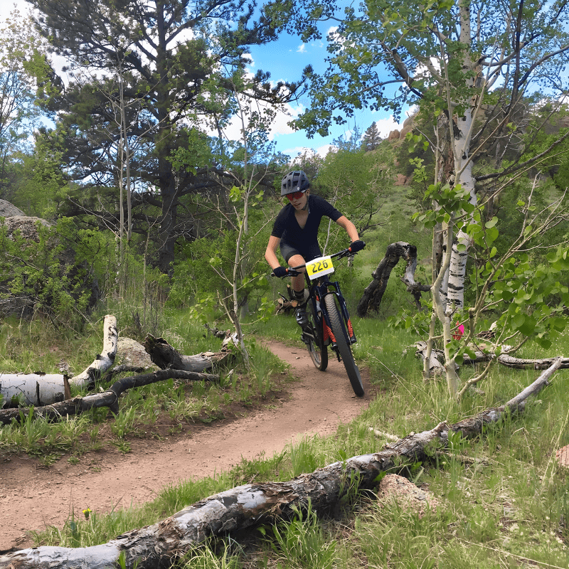 On the Stone Temple Circuit Loop trail in Curt Gowdy State Park, the image showcases a person rides a mountain bike on a narrow, winding singletrack trail through a forest. The trail is surrounded by trees and shrubs with green leaves. The mountain biker is wearing a helmet and protective gear.