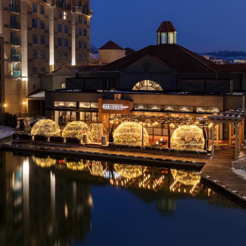 Individual dining domes illuminated with yellow-white lights in front of OAK + CRU at twilight