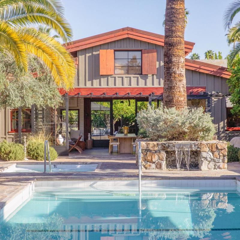A two-story Sparrows Hotel with a mix of modern and rustic design elements, featuring wood paneling and large windows, set behind a swimming pool with a small waterfall feature, surrounded by lush palm trees under a clear blue sky.