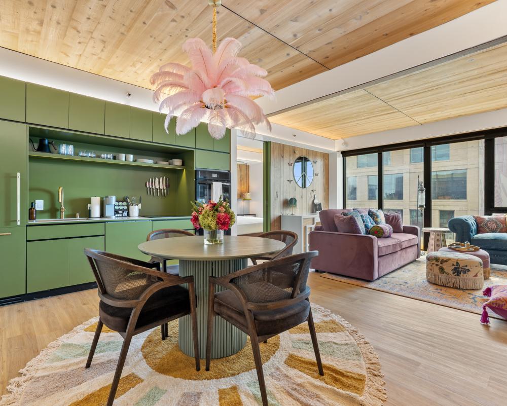 A hotel room at boheme featuring a green wall of cabinets and a kitchenette, a small dining table, and colorful couches sitting in front of a large wall of windows.