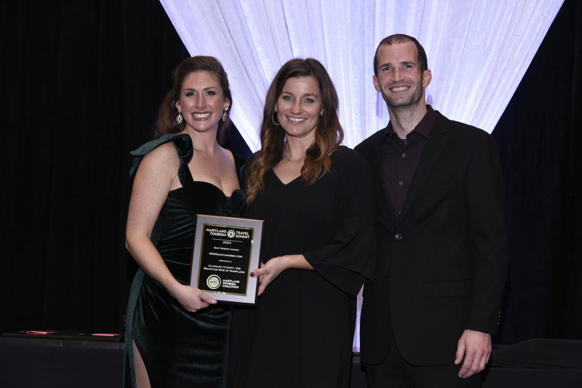 A man, and two women stand on a stage with the women sharing holding a plaque award.