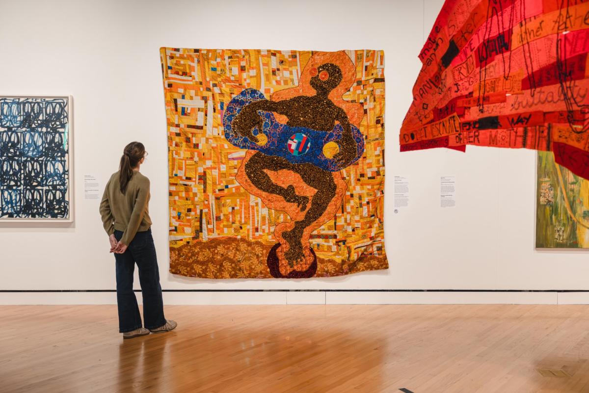 A visitor observes a vibrant textile artwork featuring abstract shapes and colors in a modern gallery space, surrounded by other art pieces.
