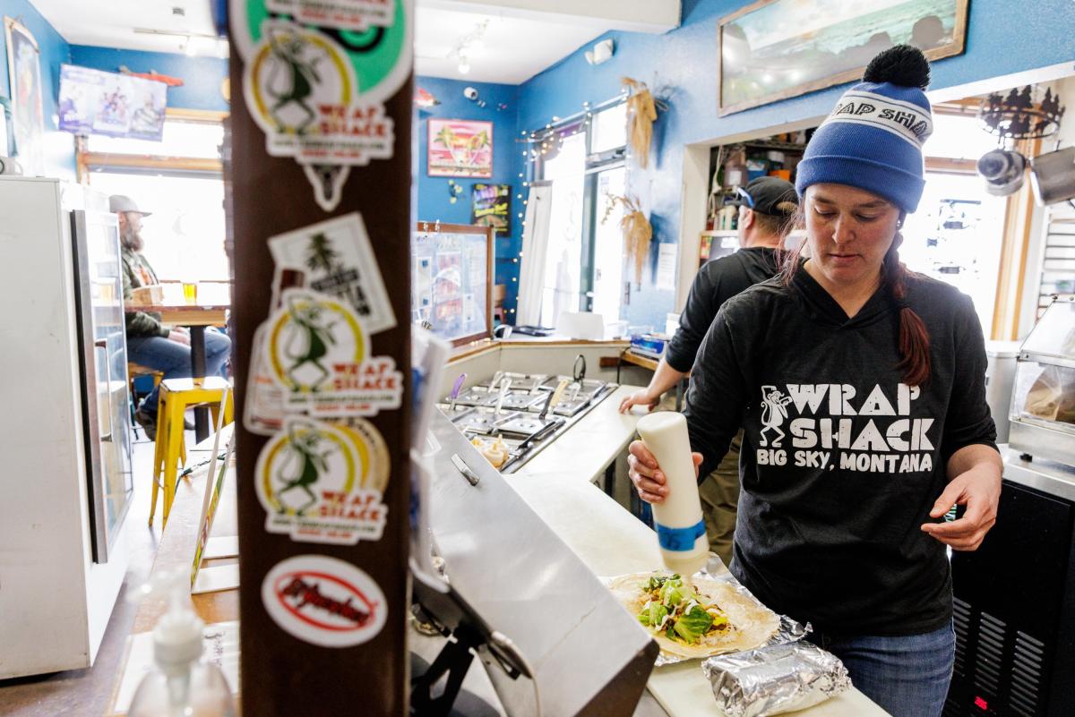 A woman in a black "Wrap Shack" shirt prepares a fresh wrap in a cozy eatery with colorful decor and patrons in the background.
