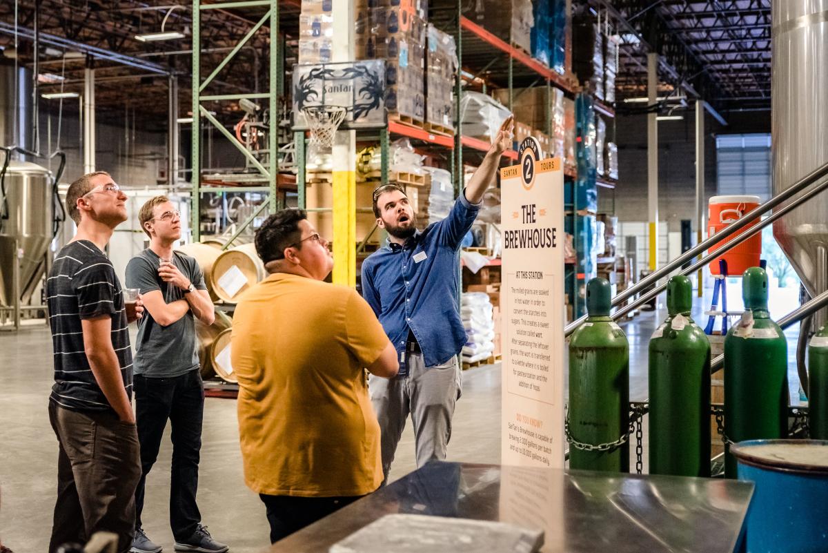 Group of 3 men touring the SanTan production site drinking sample brews with tour guide pointing up at a distiller.
