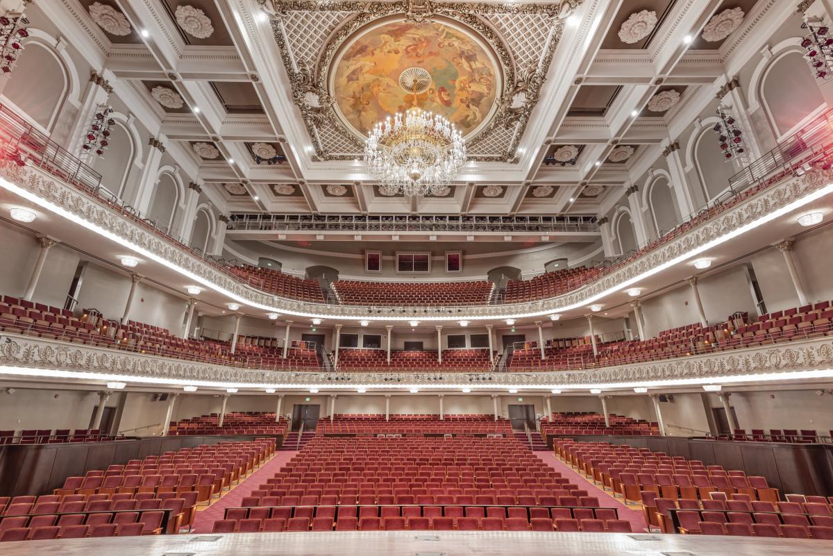 The image is of the inside of Cincinnati's Music Hall facing the audience. It shows the red seats and the goregous chandelier.