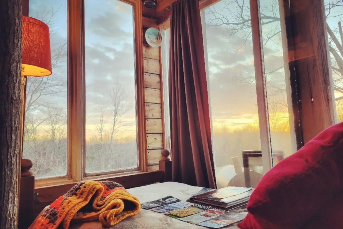 Image is of inside a rustic container with a bed surrounded by windows looking out at the sunset.