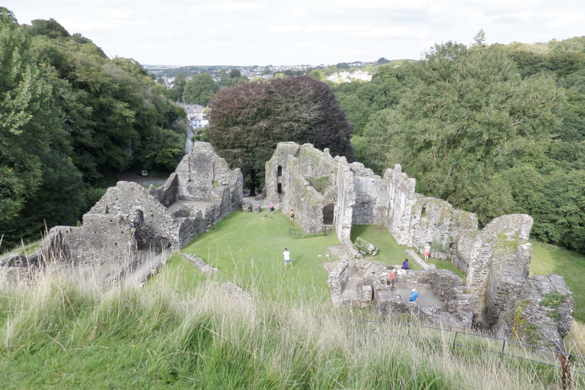 Ancient stone ruins surrounded by lush green trees and grass, with people exploring the site. The scene conveys a sense of history and tranquillity.