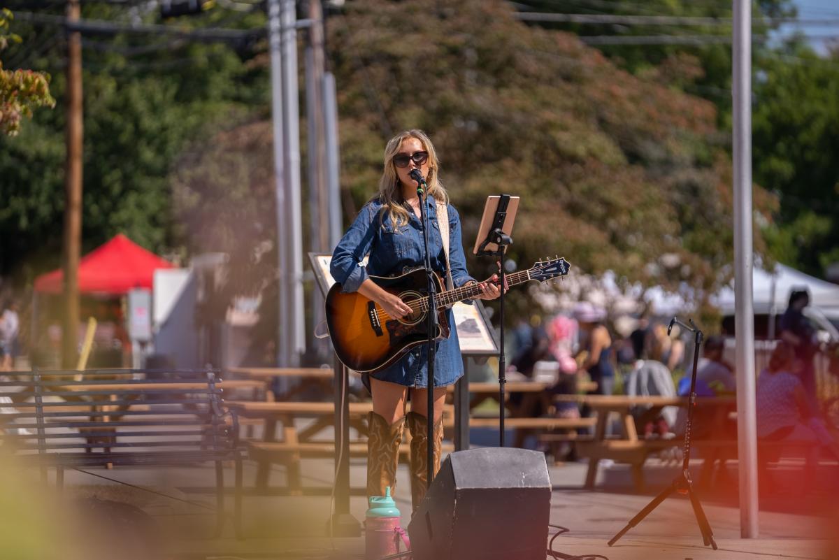 A female musician performing outdoors, playing an acoustic guitar and singing into a microphone at a local event with people and vendor booths in the background