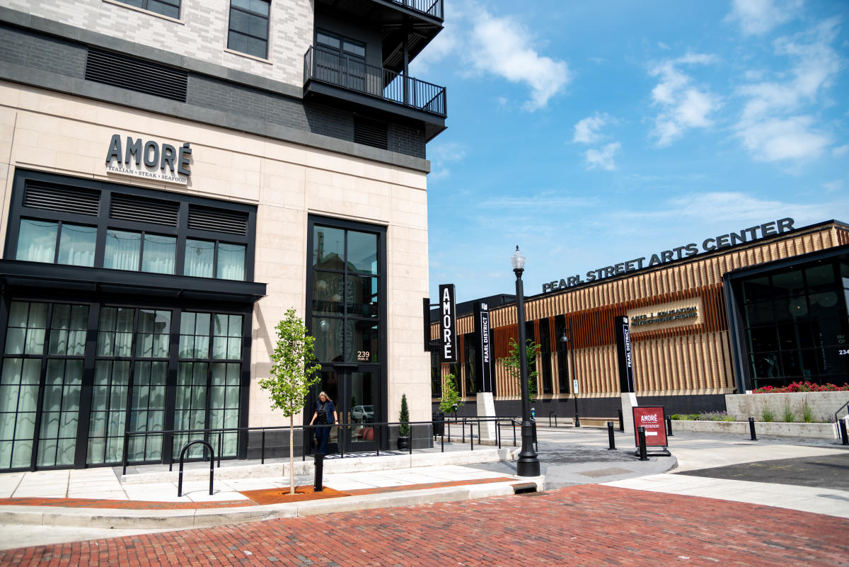 Exterior view of Amore Italian Steak Seafood restaurant and Pearl Street Arts Center in Fort Wayne’s Pearl District, with modern architecture, brick street, and clear blue sky
