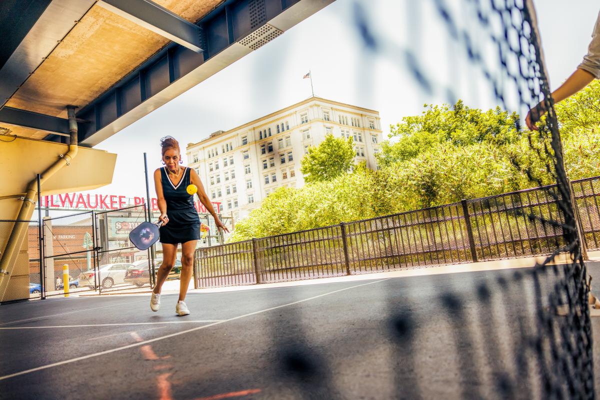 A woman playing pickleball on an outdoor court beneath an overpass in Milwaukee, focused as she returns a bright yellow ball, with a city building and greenery in the background.