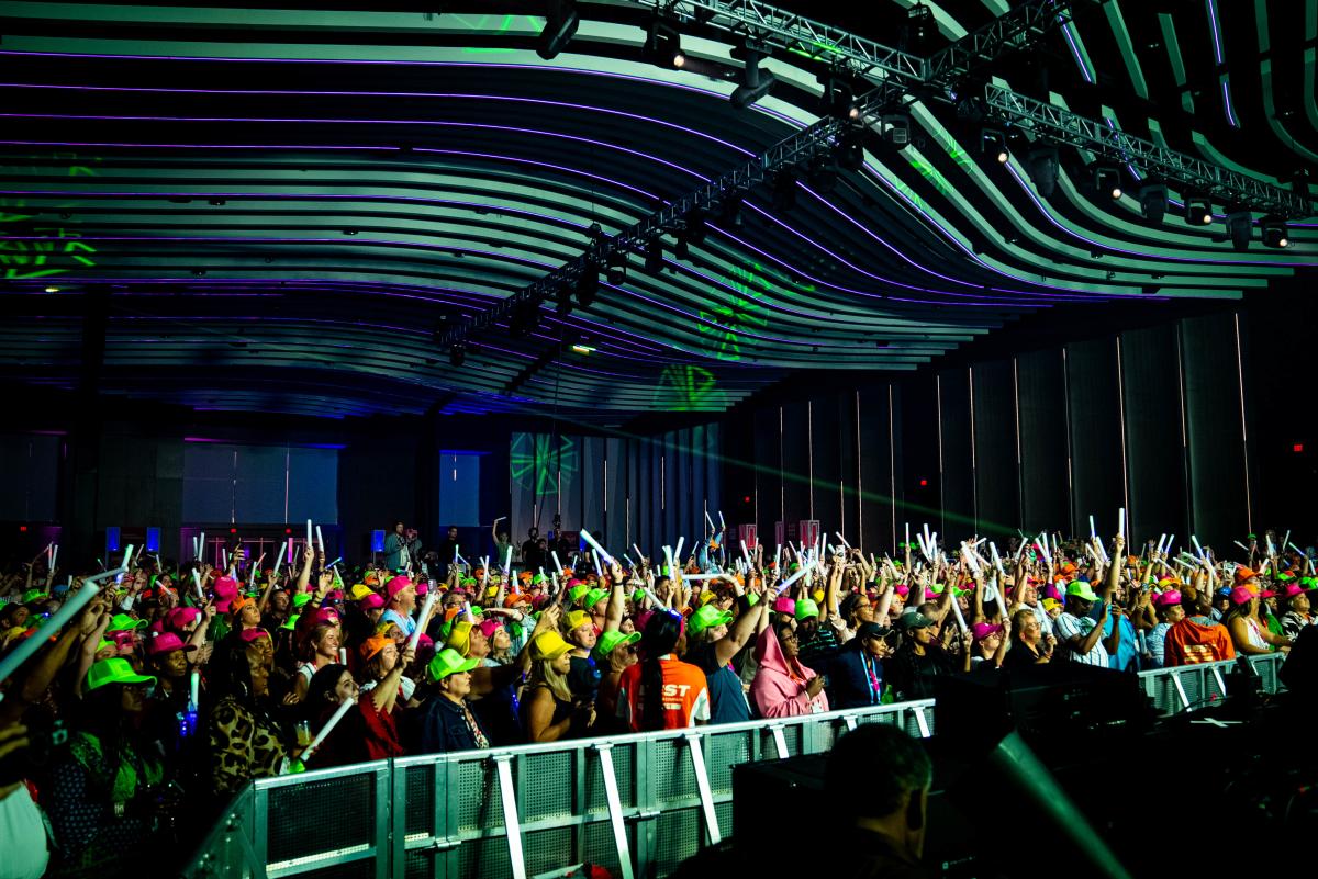 A packed crowd of energetic attendees wearing neon hats and holding light-up foam sticks at a lively indoor event. The venue features a modern, wave-like ceiling with vibrant green and purple lighting. The audience faces the stage, fully engaged in the experience, creating a festive and high-energy atmosphere.