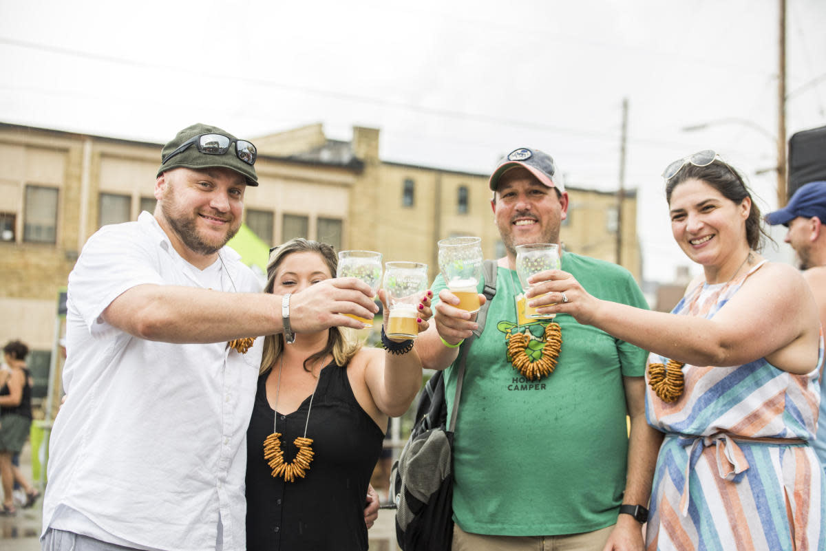 Four adults stand outdoors clinking their beer glasses together in a toast at a festival. Each person wears a pretzel necklace, a common accessory at beer events. They are smiling and appear to be enjoying themselves. The background includes a brick building and other festival attendees, with overcast skies overhead.