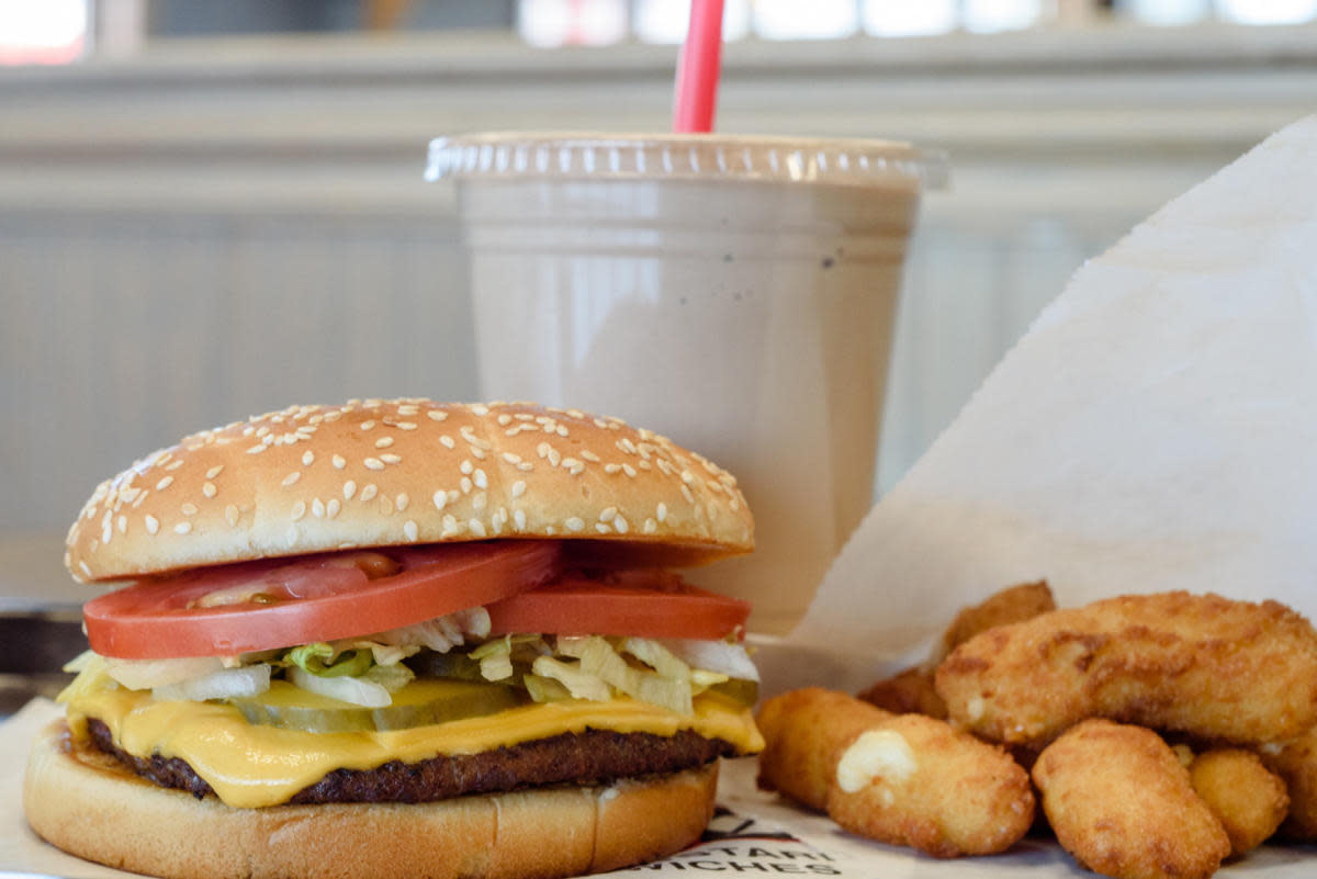 Cheeseburger with lettuce, tomato, pickles, and melted cheese on a sesame seed bun, served with cheese curds and a milkshake in the background.
