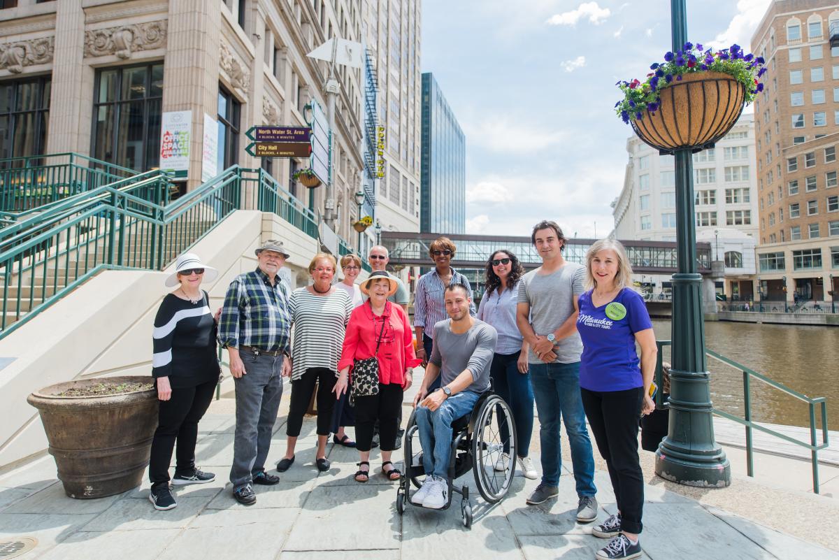 Group photo of people standing together along the Milwaukee Riverwalk on a sunny day. The group includes adults of various ages, including one person using a wheelchair. Downtown buildings, a river, a pedestrian bridge, and a lamppost with a hanging flower basket are visible in the background.