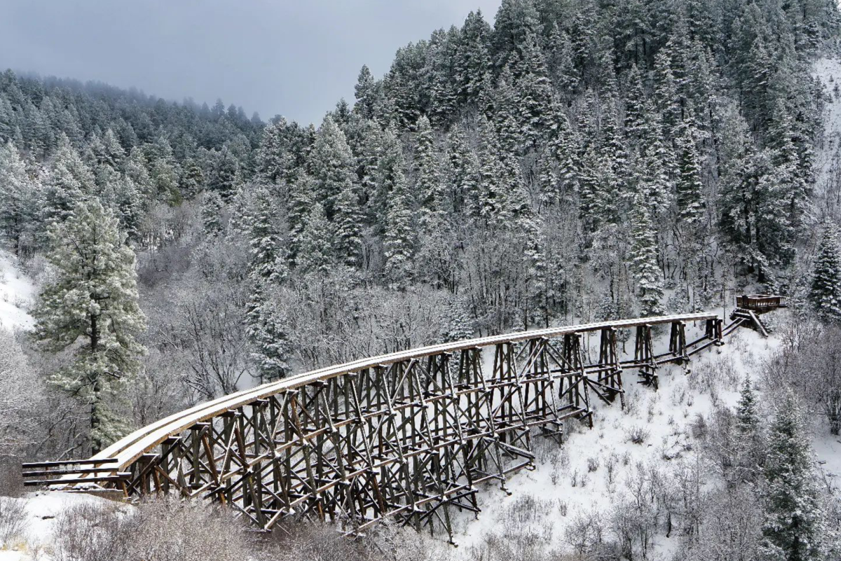 An image of a wooden trestle bridge covered in snow, extending across a forested valley filled with snow-dusted evergreen trees.