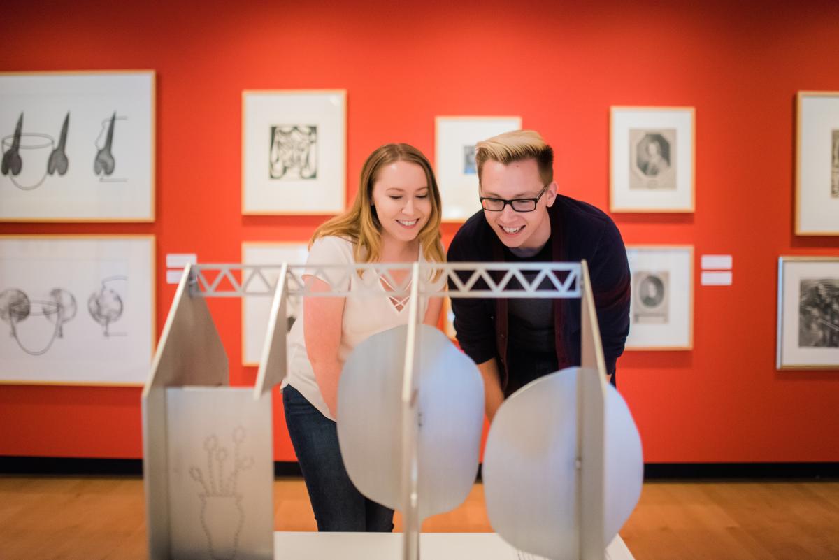 Two teenagers facing the camera looking at a sculpture shaped like a house. Behind them is a wall of more artwork.