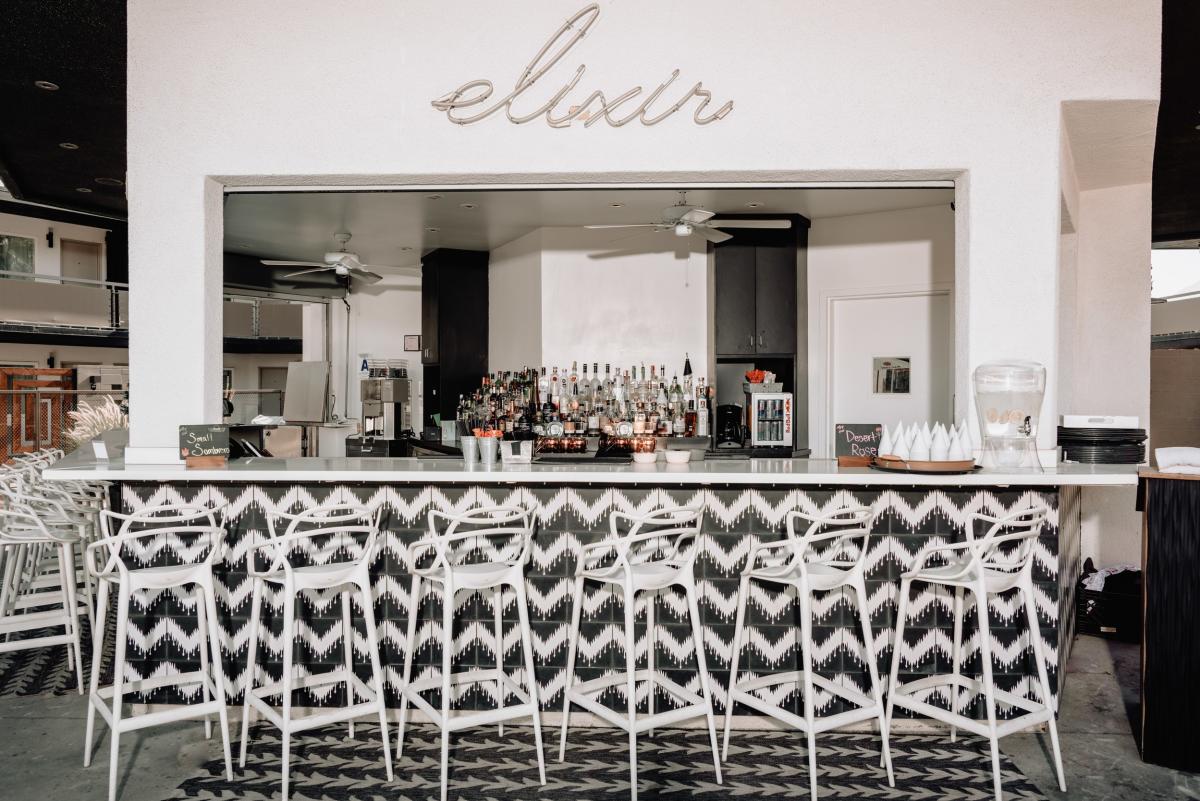 An outdoor bar counter with a stylish geometric design, white high stools, and a variety of bottles on shelves under a sign reading "elixir".