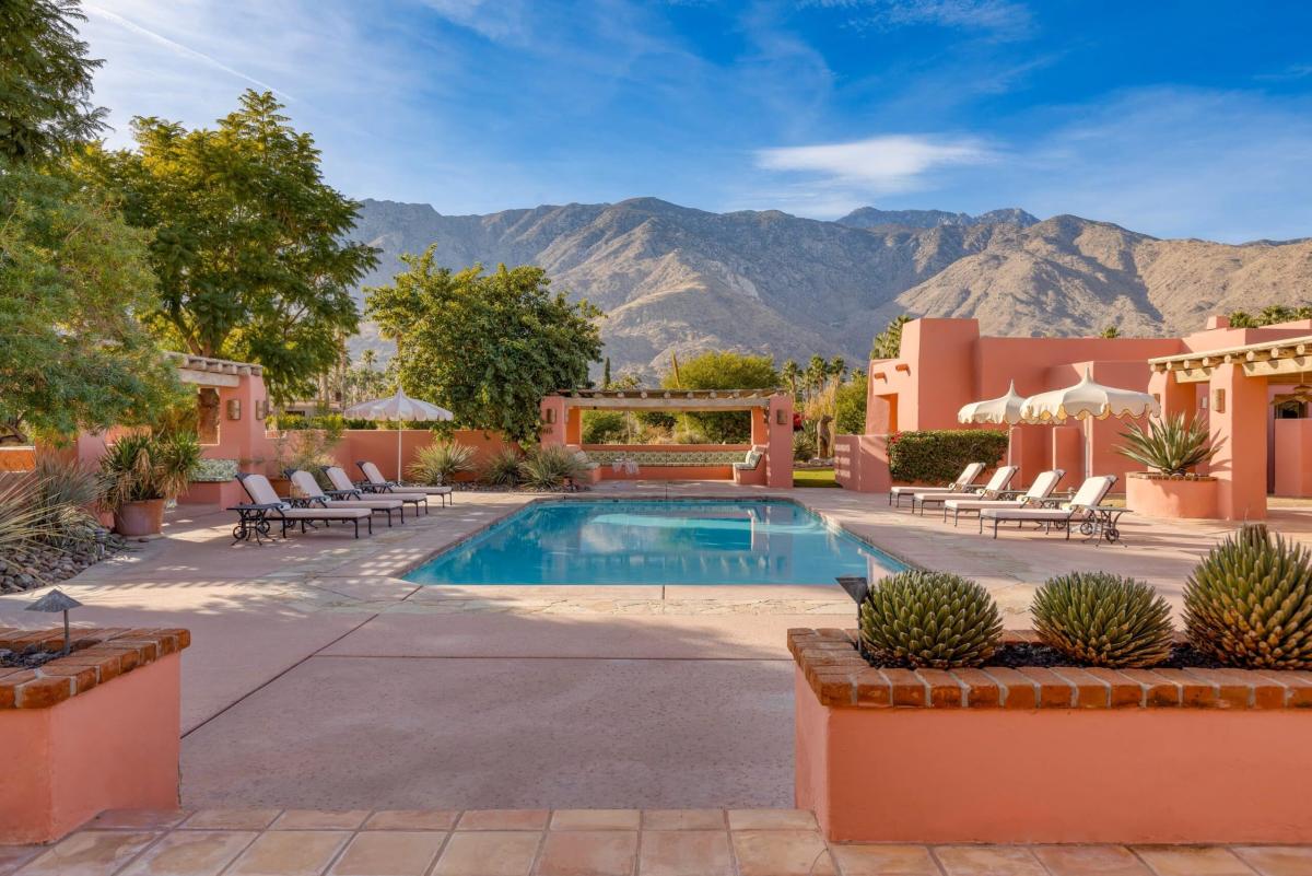 An outdoor swimming pool with surrounding sun loungers at a terracotta-colored resort, with mountains in the background and a clear blue sky above.