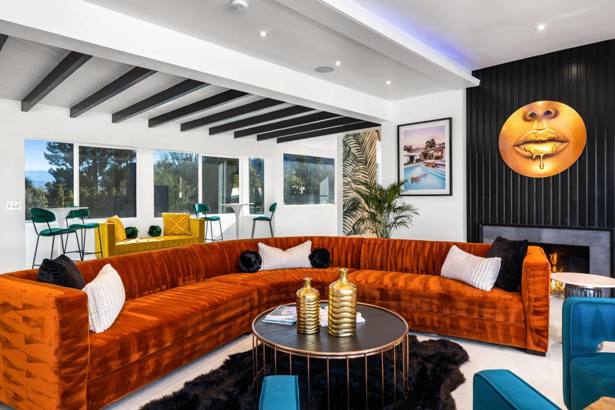 A modern living room with a large orange velvet sectional sofa, contrasting black and white throw pillows, a circular coffee table with golden vases, and a fireplace. The décor includes a dark paneled wall with a round golden artwork depicting lips, colorful chairs at a bar area by the window, and vibrant plants and framed artwork. The room features white walls with exposed gray beams on the ceiling and ample natural light.