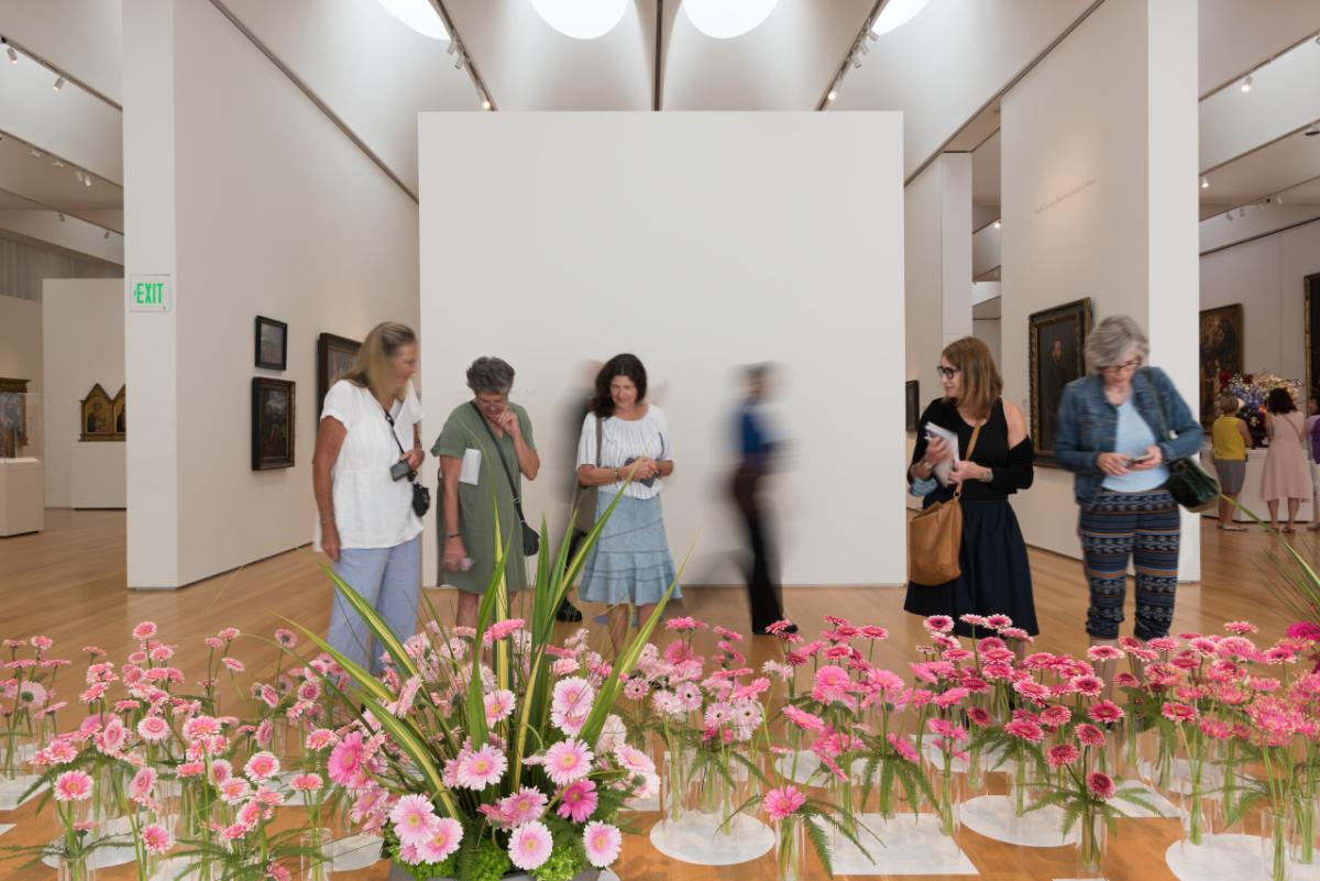 visitors inside museum gallery look at display of pink flowers in glass vases