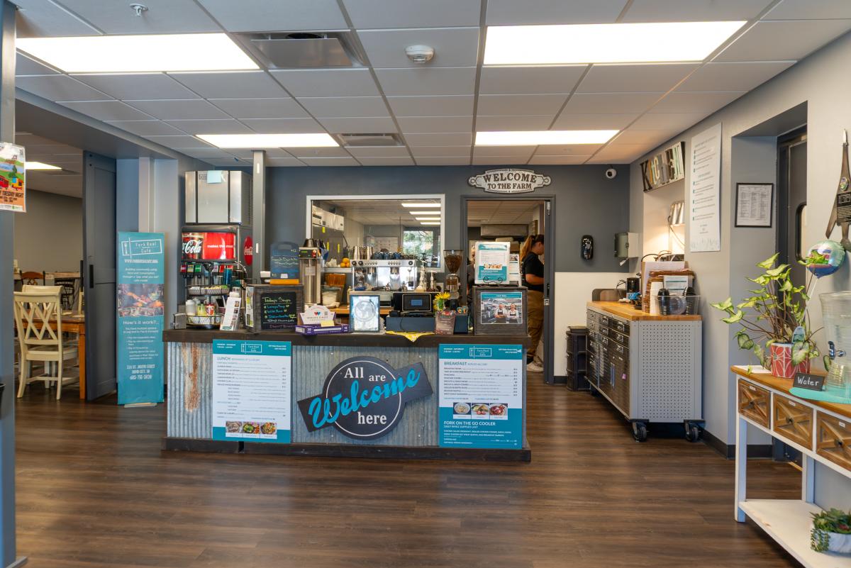 Cafe interior with a welcoming atmosphere. The counter has a sign reading "All are Welcome here." Menu boards and decor add warmth to the cozy space.