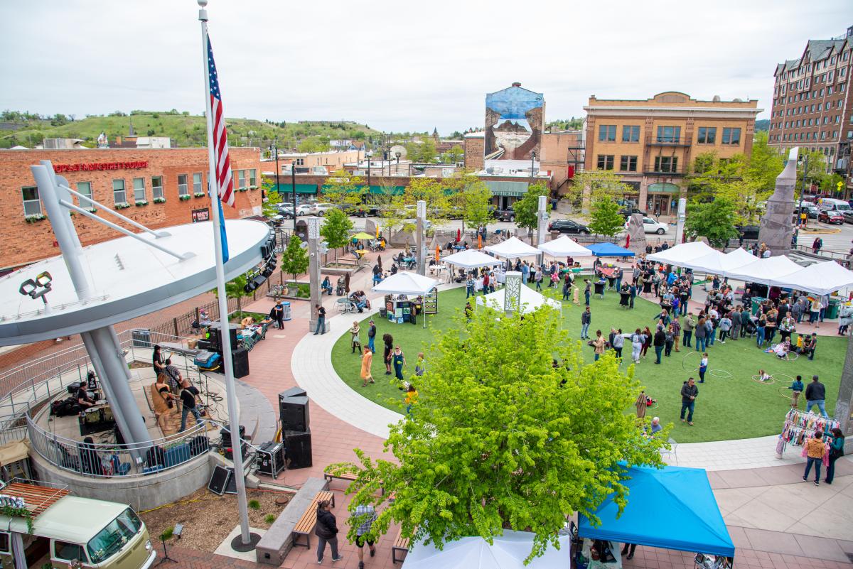 A high vantage point photo of Main Street Square while a band plays on the stage and shoppers browse the vendors in tents on the grass.