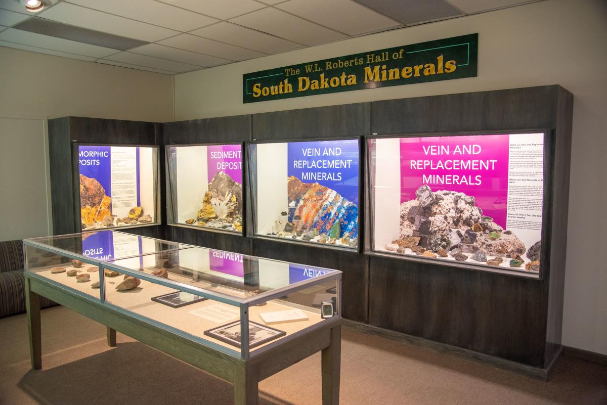 Exhibit room labeled "South Dakota Minerals" showing glass cases with colorful mineral displays and informative signs. A display table features various rock samples.