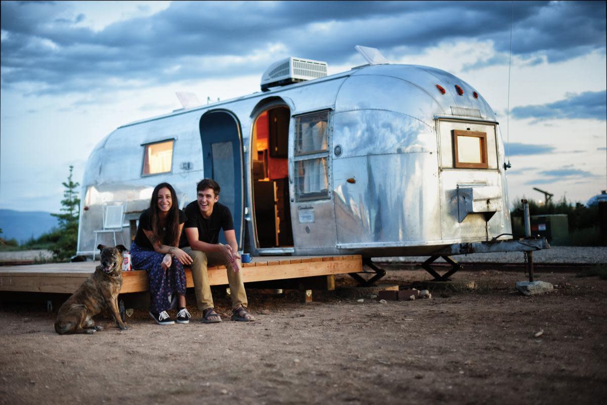 A dog, woman, and a man sitting outside of a silver vintage RV in the New Mexico desert.
