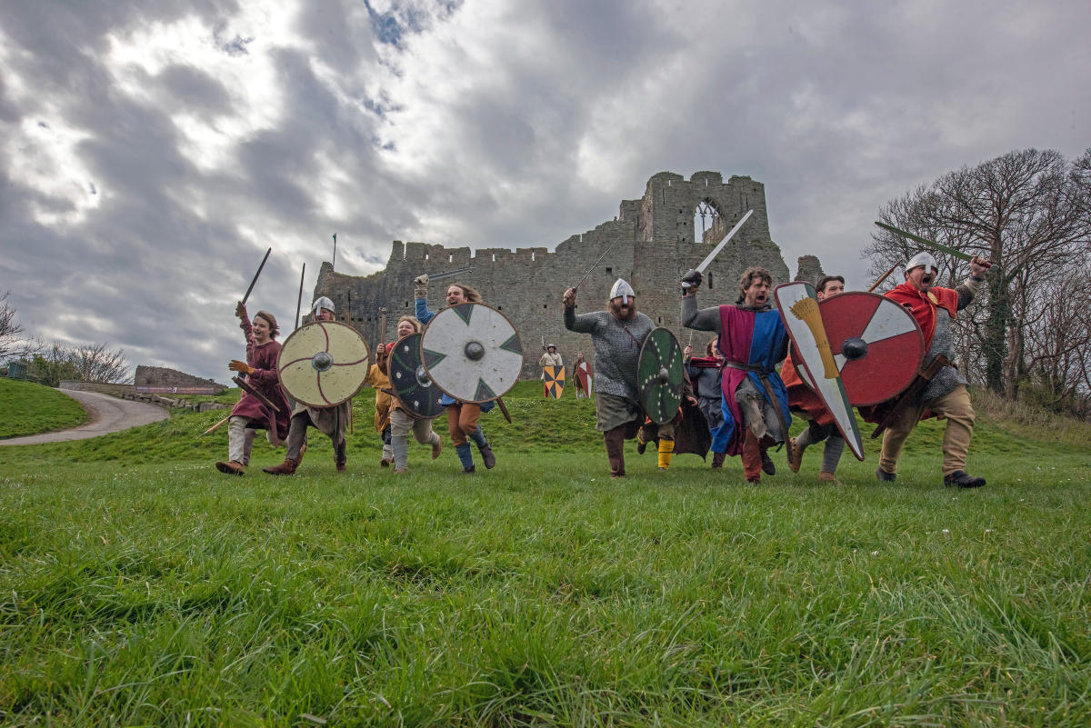 A group of people dressed as Knights charging toward the camera with Oystermough Castle in the background.