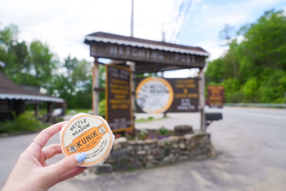 Handing holding Nettle Meadow's Kunik cheese in front of their sign