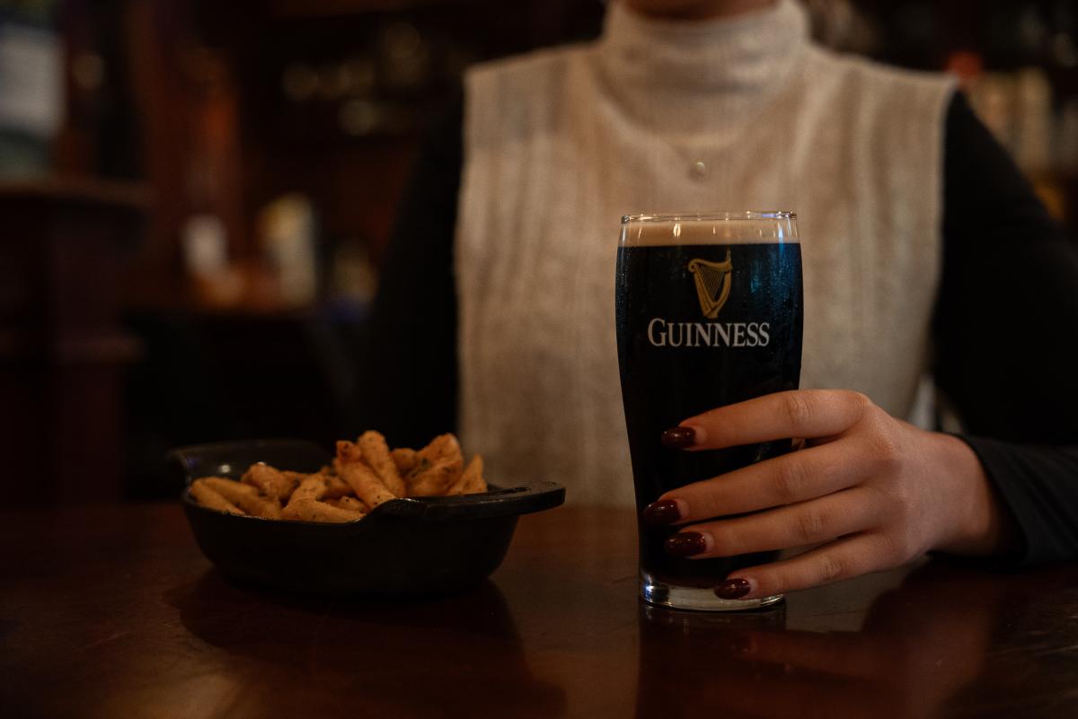 A young woman (Brooklyn Newell, face out of frame) holds a glass of Guinness beer at a table at The Goose's Acre. Beside the glass is a small skillet of seasoned french fries.