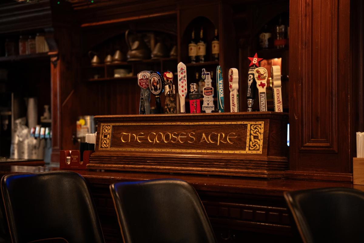 The bar at The Goose's Acre. It features a sturdy wood placard with the business name engraved in gold, surrounded by Celtic patterns. Behind this placard are the levers for eleven beers on tap. Among them are Union Jack IPA, KONA, and Saint Arnold.